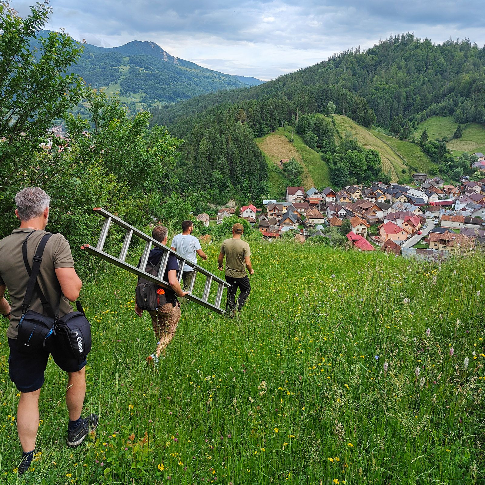 A group of people hiking on an hill with view on a village whilst carrying a batch of repellers, battery cases and a ladder.